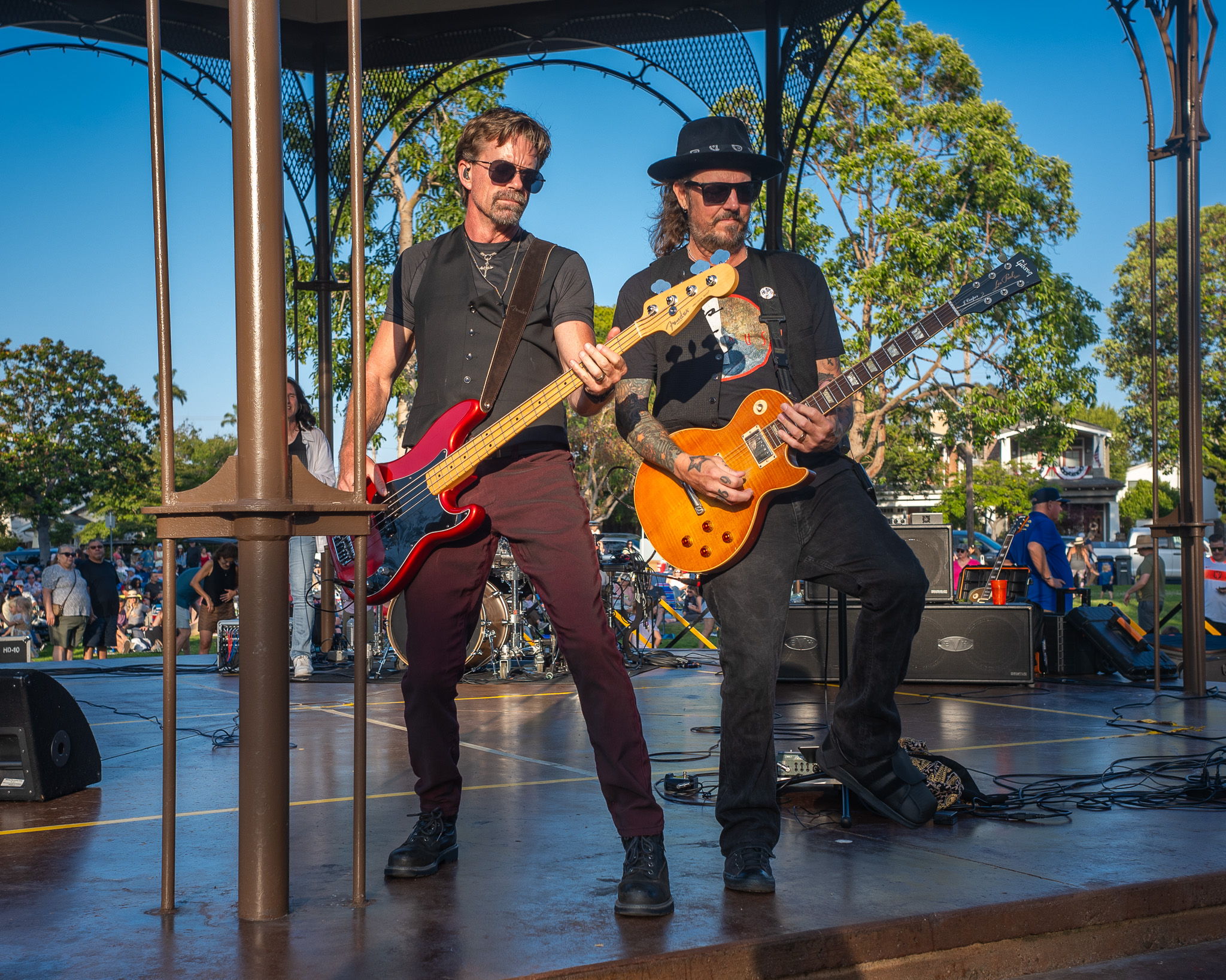 Two guitarists performing under an outdoor bandstand — one on bass guitar in a grey vest, the other playing a sunburst Gibson Les Paul, wearing a black hat and sunglasses.