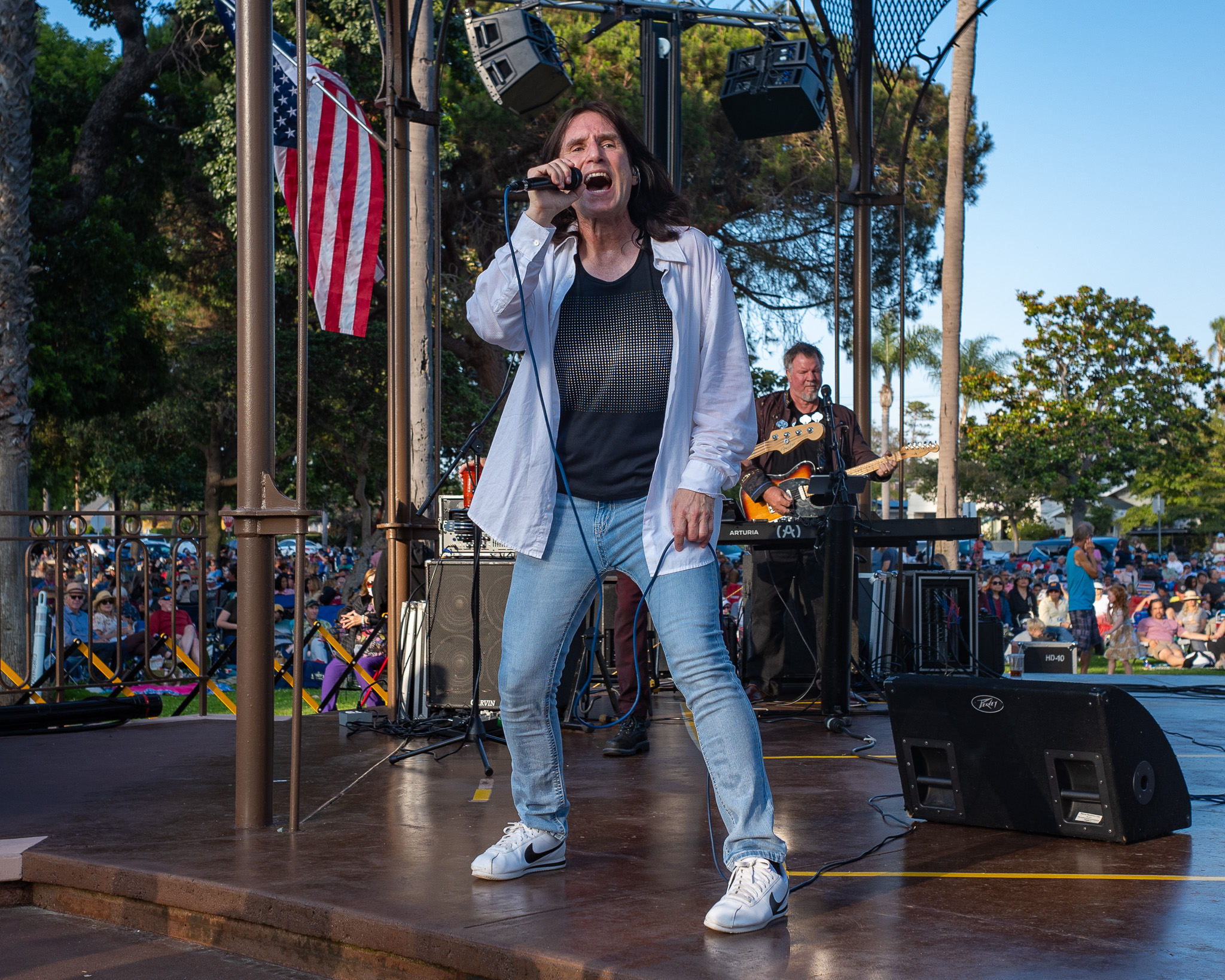 A singer in a white shirt belts into a microphone on an outdoor stage, with a bassist performing behind him.
